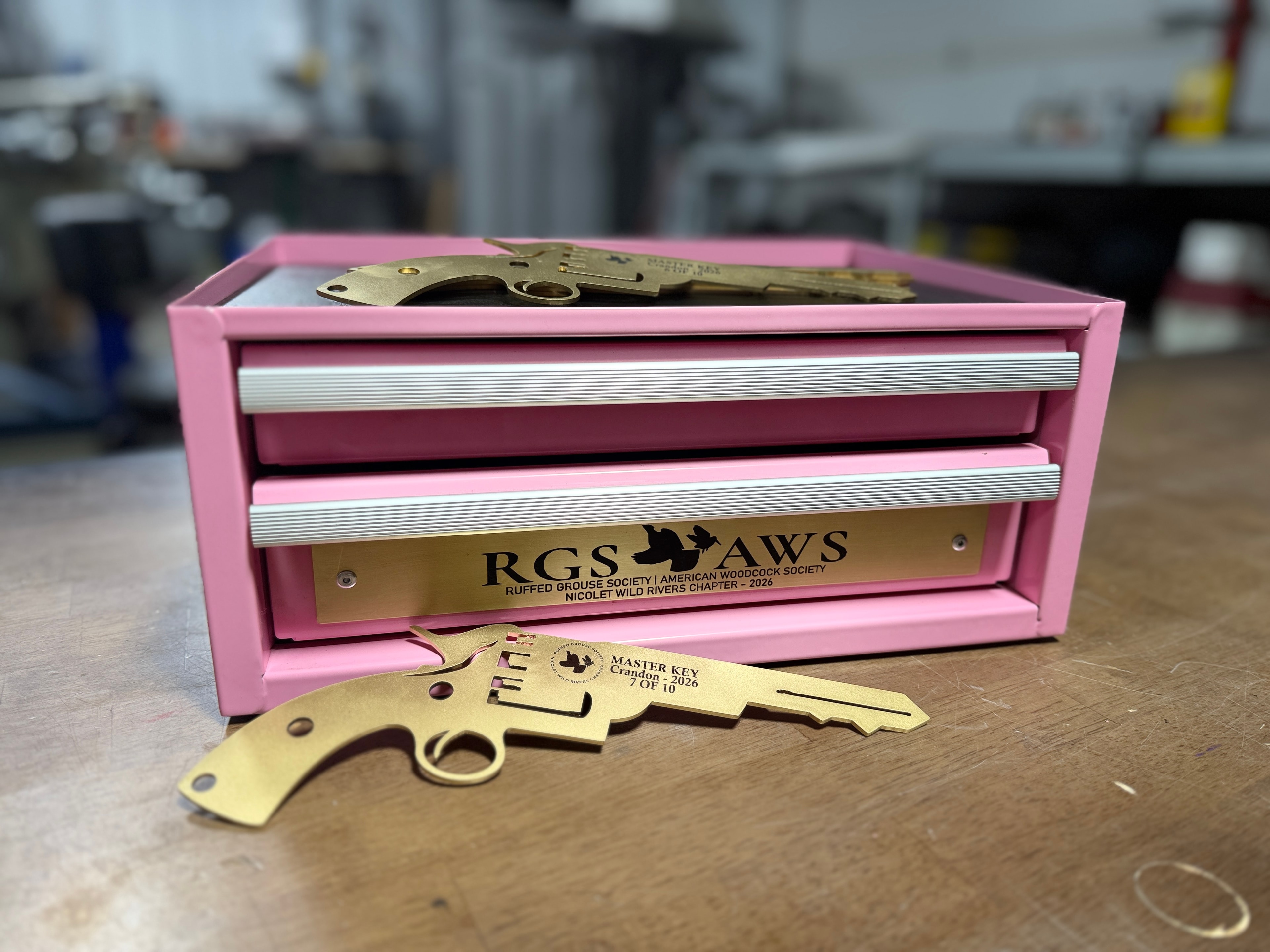Laser-engraved pink mini toolbox with RGS & AWS Ruffed Grouse Society / American Woodcock Society Nicolet Wild Rivers Chapter 2026 brass plate, and brass revolver-shaped master key — by Northwoods Firearms